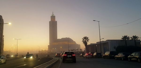 Traffic on road by buildings against sky during sunset