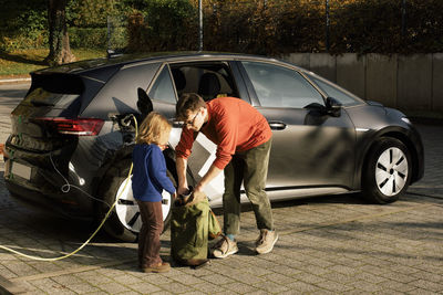 Father searching in backpack with daughter standing near electric car being charged at station