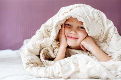Portrait of smiling boy hiding under blanket in bed at home