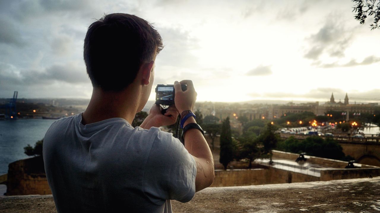 REAR VIEW OF MAN PHOTOGRAPHING RIVER AGAINST CITYSCAPE