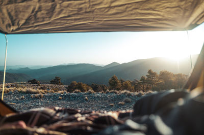 View through tent on hilly scenery lightened by bright rays of sun rising on horizon on blue sky in early morning