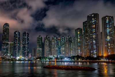 Illuminated modern buildings by river against sky at night