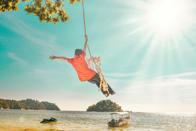 Low angle view of boy swinging on rope swing at beach against sky