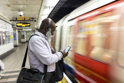 Bald senior businessman using smart phone near speeding train at station