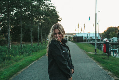 Portrait of smiling young woman standing by road against trees