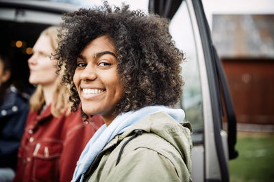 Portrait of smiling young woman with curly hair standing with friend by car