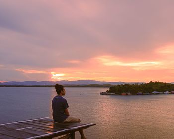 Rear view of man sitting on lake against sky during sunset