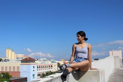 Young woman sitting on retaining wall at building terrace against blue sky during sunny day