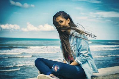 Portrait of woman with tousled hair sitting on retaining wall by sea against sky