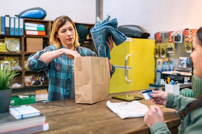 Woman cashier putting jeans into paper bag while female customer holding credit card to pay in store