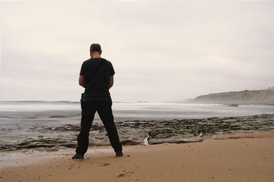 Rear view of man standing at beach against sky during sunset