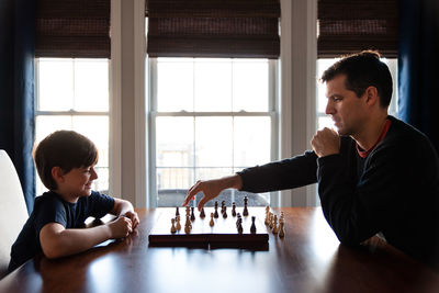 Father and son sitting at a table indoors playing a game of chess.