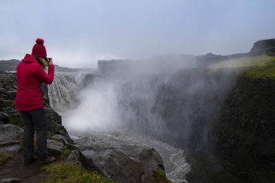 Woman photographing while standing by waterfall against sky