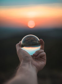 Person holding crystal ball against sky during sunset