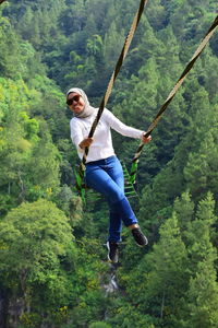 High angle portrait of cheerful woman sitting on swing against forest