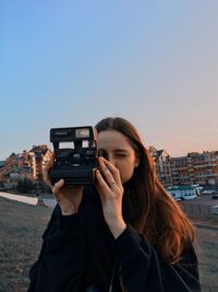 Young woman photographing camera against sky in city