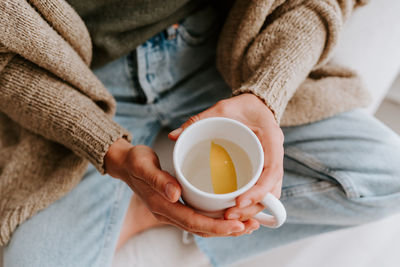 From above of anonymous female sitting with cup of water with slice of fresh lemon in morning at home