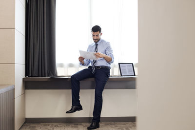 Full length of businessman reading documents while sitting on window sill in hotel room