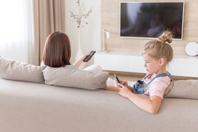 Girl using smart phone sitting by mother on sofa at home