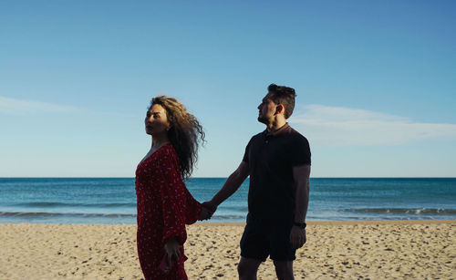 Young woman with man standing at beach against clear sky with holding hands