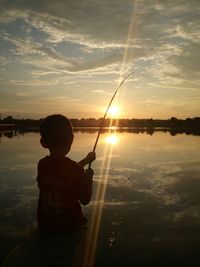 Boy fishing in lake against sky during sunset