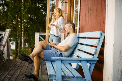 Smiling man sitting on bench while woman talking on mobile phone at porch