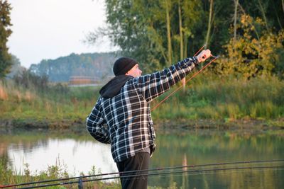 Man using slingshot while standing by fishing rod at lakeshore against trees