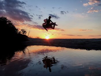 Silhouette woman jumping over pond against sky during sunset