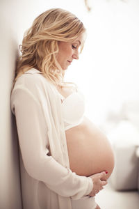 Side view of pregnant woman touching abdomen while leaning on wall at home