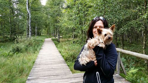 Portrait of smiling mature woman holding dog on boardwalk in forest