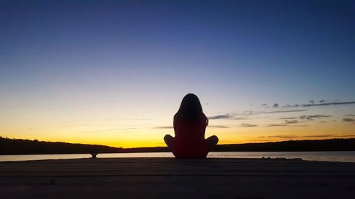 Silhouette woman sitting by sea against clear sky during sunset