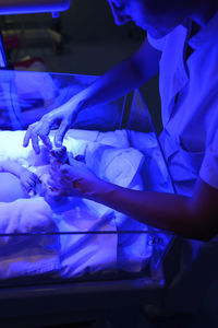 From above crop person holding small foot of sick tiny baby lying in infant incubator in icu under ultraviolet for phototherapy in hospital