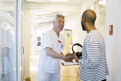 Senior doctor with clipboard greeting young man at hospital corridor