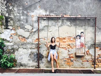 Portrait of happy woman sitting on swing against wall