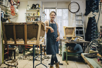 Portrait of mature male craftsperson standing by incomplete sofa in upholstery workshop