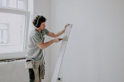 Young caucasian male plasterer listens to music through headphones and cleans the putty trowel