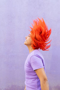 Side view of excited adult female with closed eyes shaking bright ginger dyed hair against violet wall on street