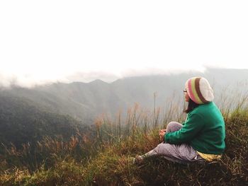 Woman relaxing on grassy field at mountains against sky during foggy weather