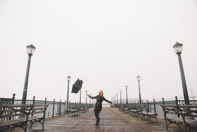 Full length of young woman holding umbrella while standing on pier against sky