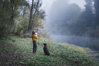 A woman is holding a baby and looking at a dog near a river