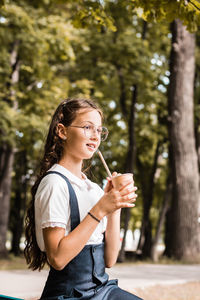 A pupil in eyeglasses drinks a beverage from an eco paper cup with a straw in the park