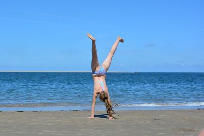 Full length of girl practicing handstand at beach against sky during summer