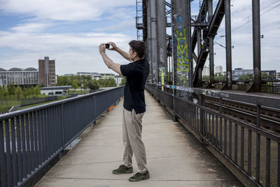 Young man photographing through mobile phone while standing on bridge