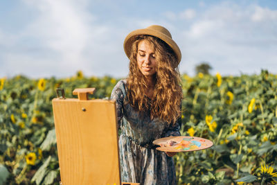 A young woman with curly hair and wearing a hat is painting in nature. 