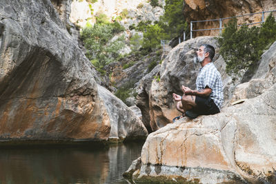 Man meditating on top of a rock at the foot of a mountain river