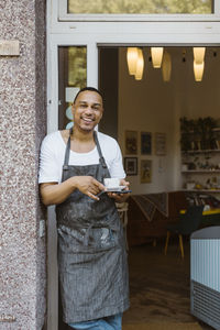 Smiling male owner with coffee cup leaning on wall near cafe doorway