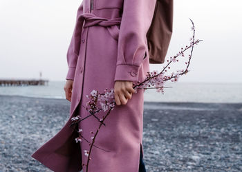 Cropped unrecognizable female in pink coat standing with blooming cheery flower twig