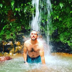 Portrait of shirtless man standing below waterfall