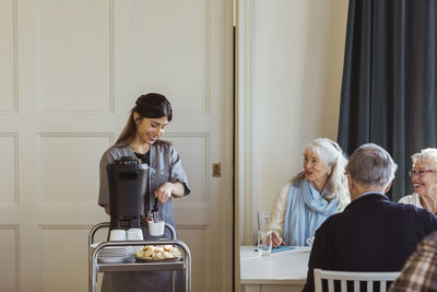 Smiling female healthcare worker serving coffee to senior women and man at retirement home