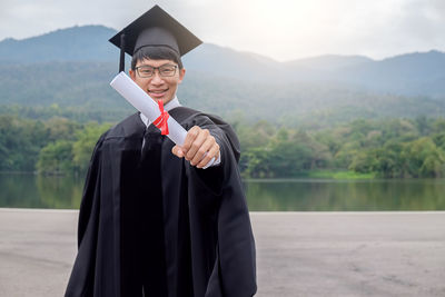 Portrait of happy young man in graduation gown standing against mountain